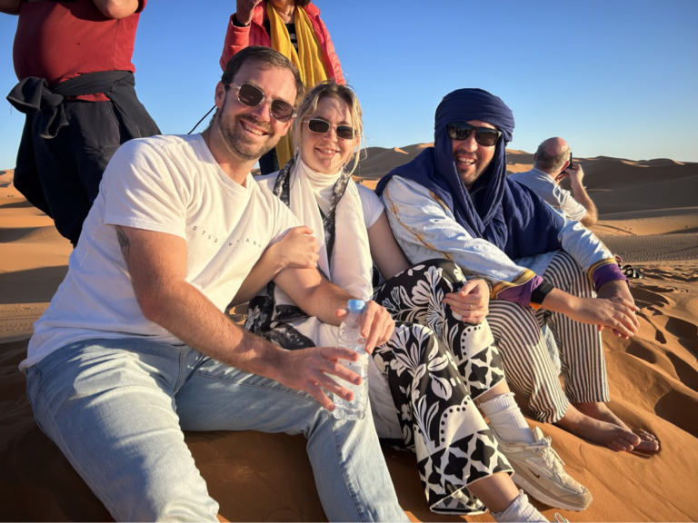 A couple poses with local tour guide Omar Imerhan on a sand dune in the Sahara desert, giving a glimpse into what trekking during a romantic trip to Morocco may entail.
