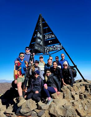 A group of trekkers poses proudly at the summit of Mount Toubkal beneath the iconic iron pyramid marker, framed by a brilliant blue sky.