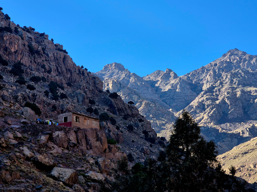 A rustic house clings to the mountainside in Morocco’s High Atlas range, framed by jagged peaks and a deep blue sky during a clear day trip.