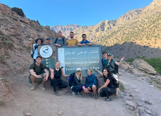 A smiling group of hikers poses at the Toubkal National Park entrance sign, surrounded by rugged mountain peaks under a bright blue sky in Morocco.
