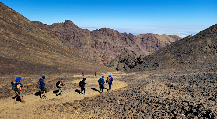 A group of hikers treks along a winding path through Morocco’s rocky High Atlas Mountains, surrounded by rugged peaks and a clear blue sky.