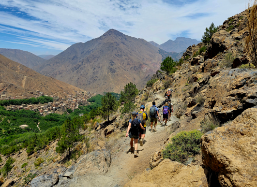 A group of hikers treks along a narrow mountain path above a lush valley in the High Atlas Mountains, surrounded by rugged peaks and scattered pine trees.