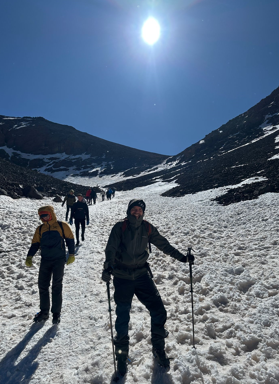 Hikers bundled in winter gear ascend a snow-covered trail on Mount Toubkal under a bright sun, with steep peaks flanking the icy path.