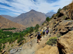 A group of hikers walks along a narrow mountain trail overlooking a green valley and a village below.