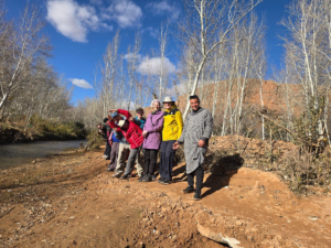 A group of people standing together on a dirt trail beside a river, surrounded by leafless trees under a bright blue sky.