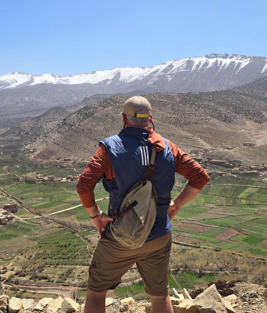 A man stands on a rocky overlook with hands on hips, gazing across a green valley toward snow-capped mountains under a clear blue sky.