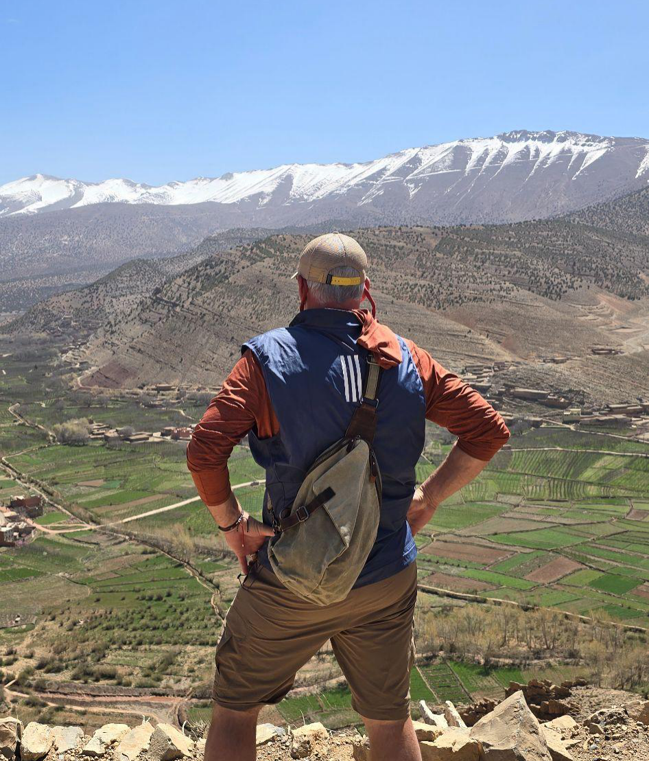 A man stands on a rocky overlook with hands on hips, gazing across a green valley toward snow-capped mountains under a clear blue sky.
