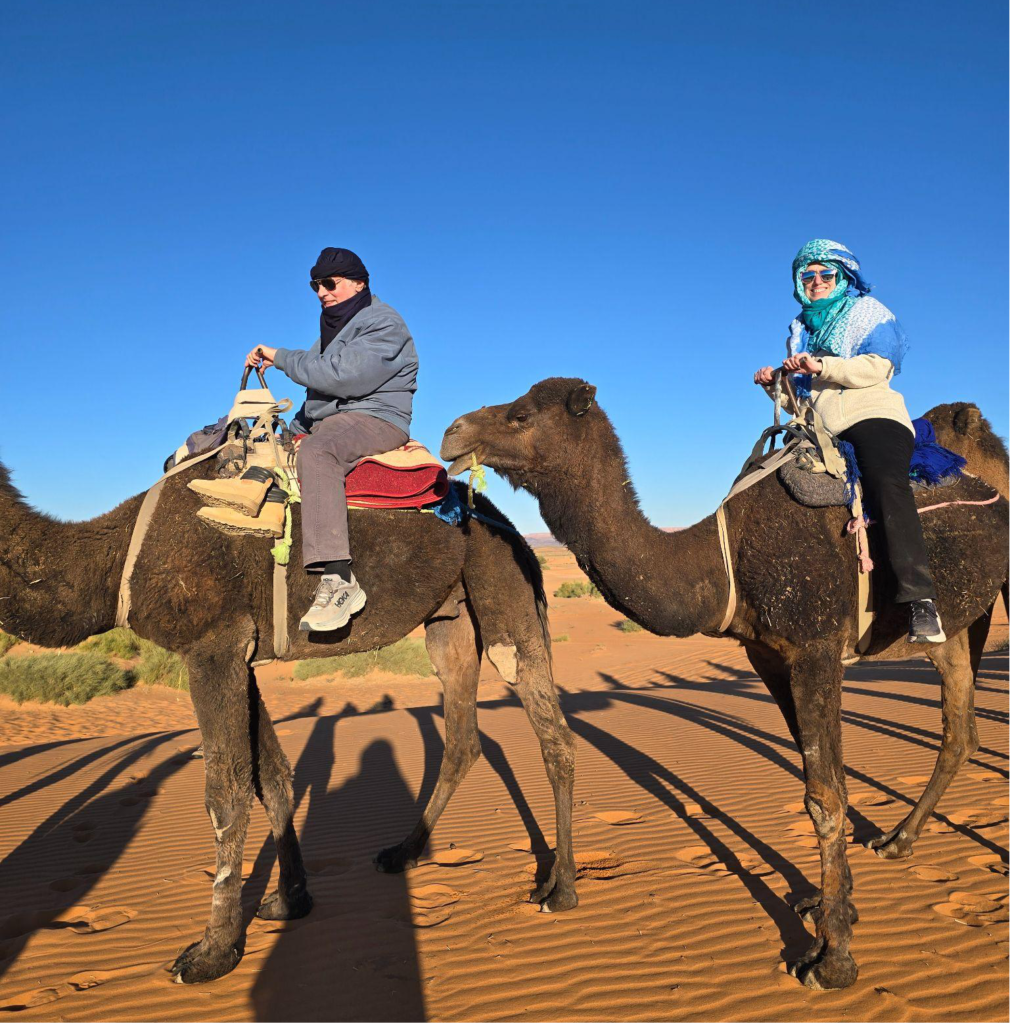 Two tourists ride camels across golden desert dunes under a clear blue sky.