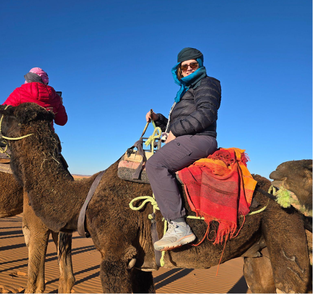A bundled-up traveler rides a camel across sunlit desert dunes under a clear blue sky.
