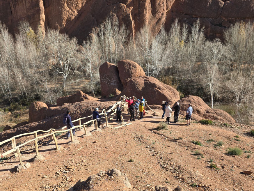 Group of travelers walking down a rocky path with large red rock formations and leafless trees in the background.
