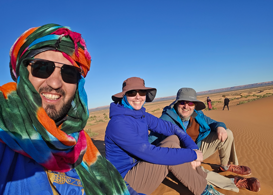 Three friends dressed for the desert pose on a sunlit dune with sweeping views of the Moroccan Sahara behind them.